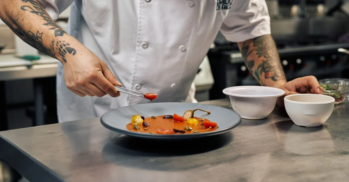 Un chef tatoué décore un plat gastronomique dans une cuisine professionnelle.