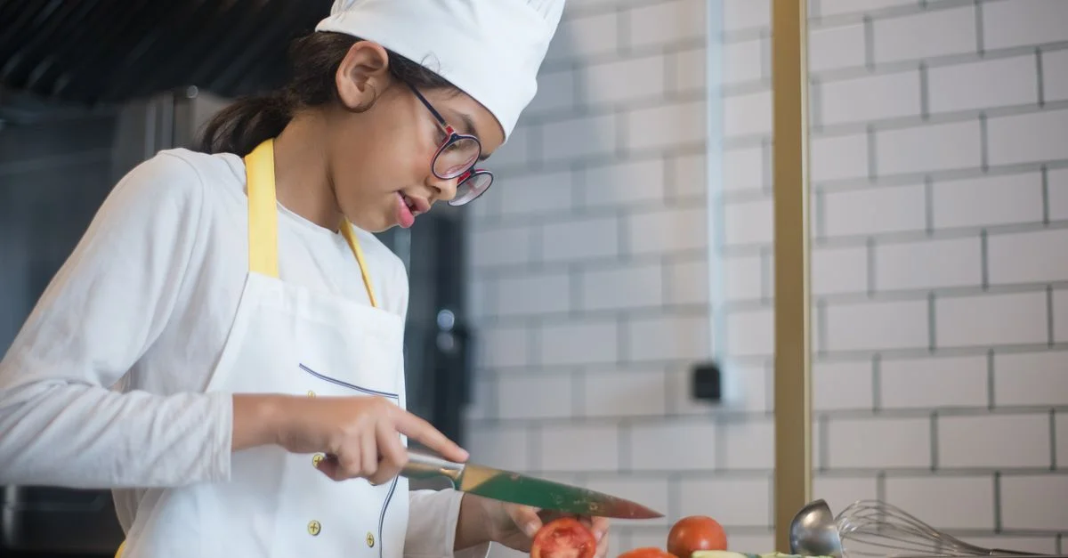 Une jeune fille portant un chapeau de chef et des lunettes coupe des légumes dans une cuisine moderne.