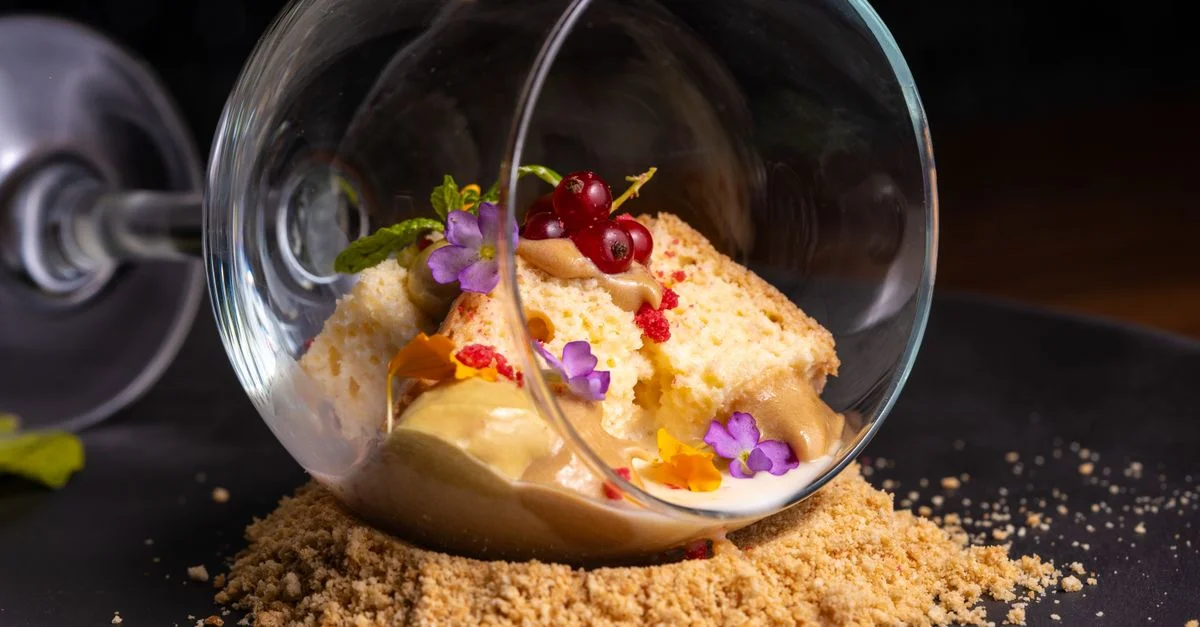 Dessert innovant servi dans un verre à l'envers avec des garnitures colorées sur une assiette noire.